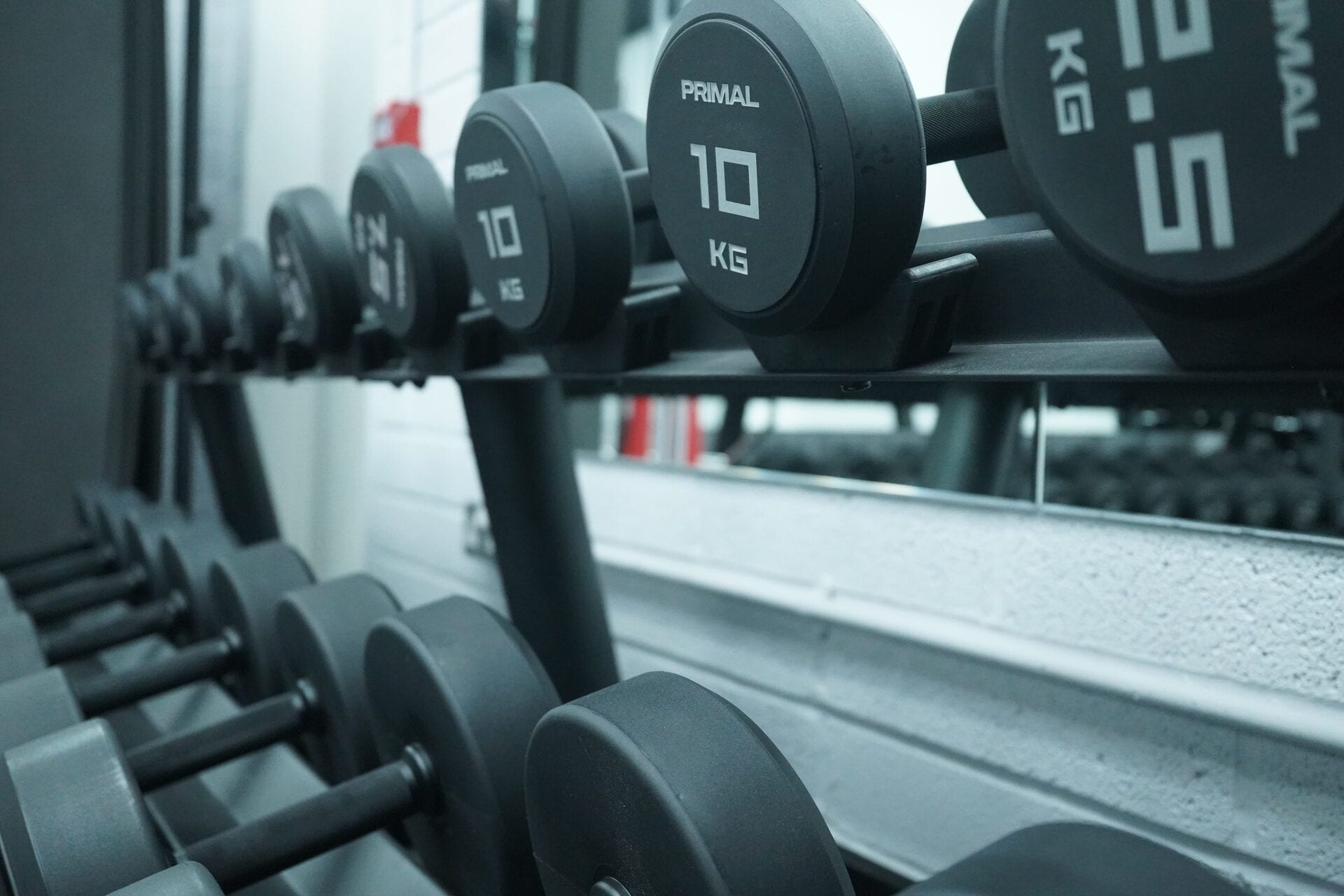 Tiered rack of PRIMAL rubber dumbbells against the white brick wall and mirror at CXHW's Hackbridge coaching studio